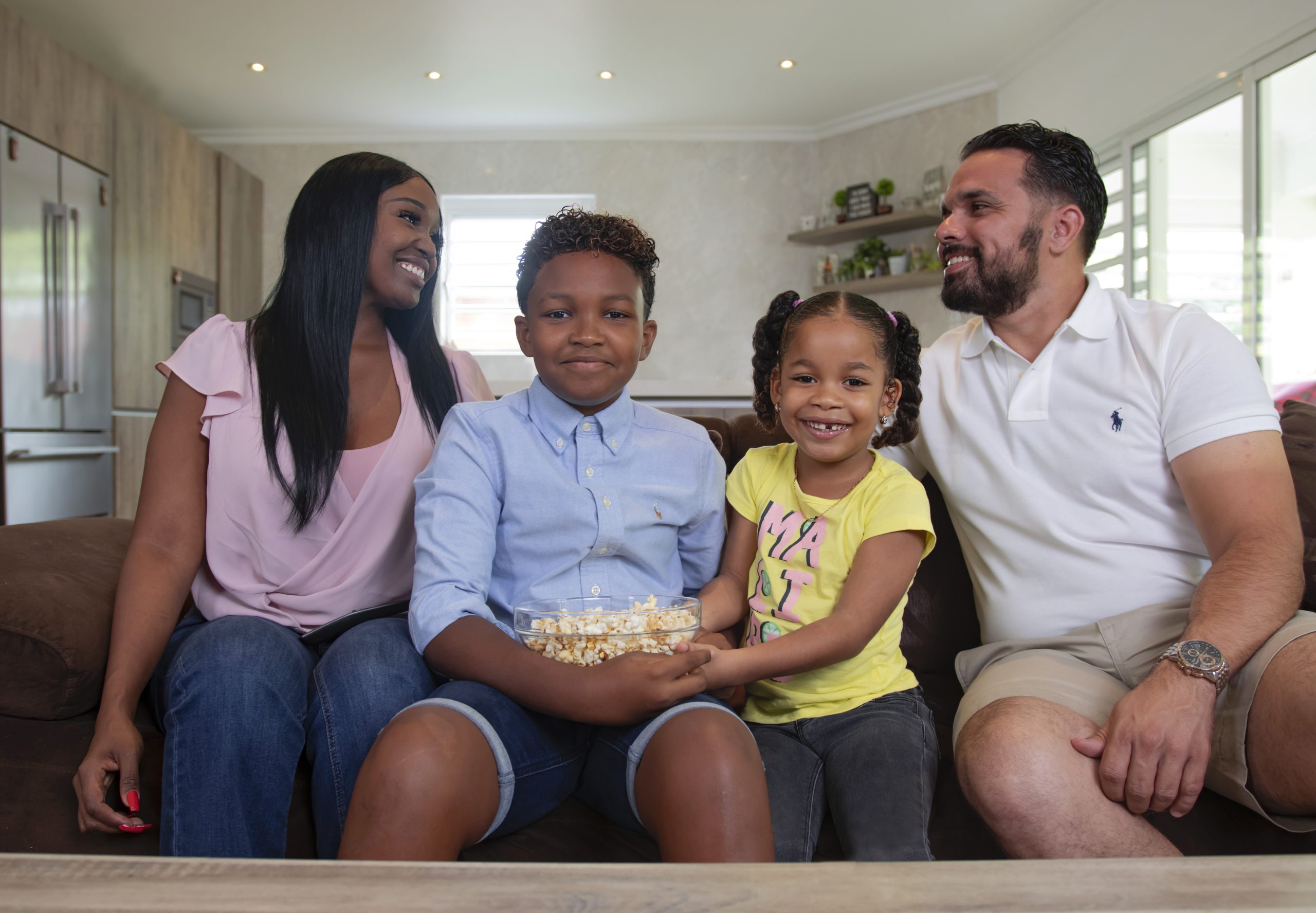 Man, woman, son and daughter are sitting on the couch. The son holds a bowl of popcorn