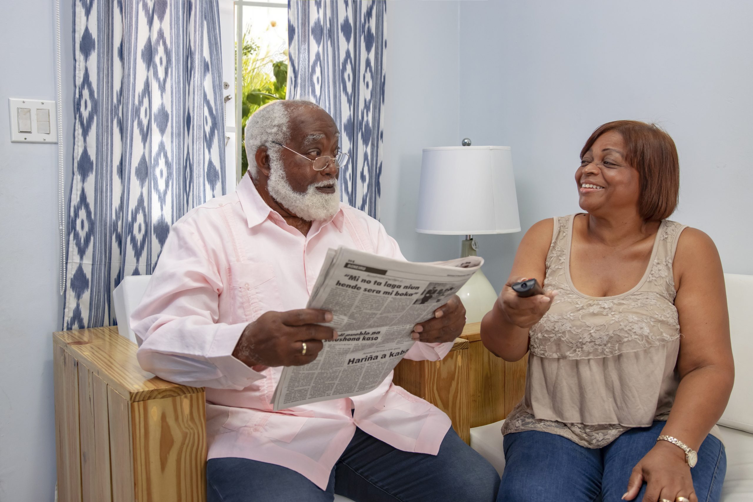 Man and woman are sitting on a bench, the woman with a tv remote in her hand and the man is reading a paper